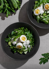 Green summer salad with arugula, cucumbers and egg on black plate. Top view food with Sunny light.