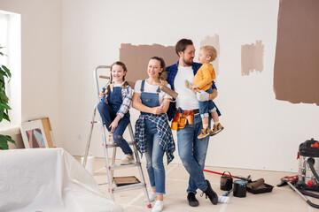 A very stylishly dressed family of four is standing in the apartment, which is being renovated. A very cute 8-year-old girl is sitting on a ladder and holding a paint roller in her hand.