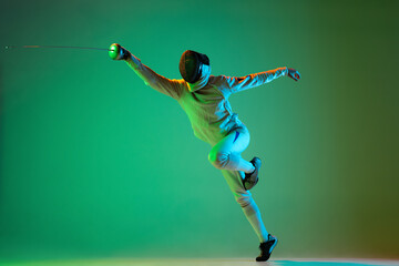 Wall Mural - Energetic young man, fencer with smallsword practicing fencing isolated on green background in neon light. Sport, energy, skills, achievements