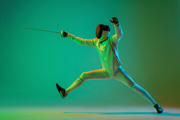 Wall Mural - Studio shot of young man, fencer with smallsword practicing fencing isolated on green background in neon light. Sport, energy, skills, achievements