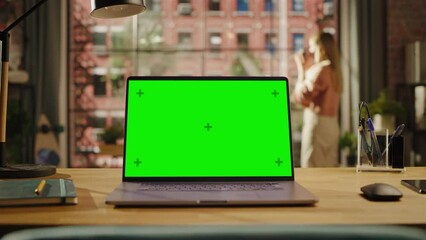 Fototapete - Laptop Computer Display with Mock Up Green Screen on a Table in Living Room. Chroma Key Monitor at Home or Creative Loft Office with Female Drinking from a Mug and Looking Out of Window in Background.
