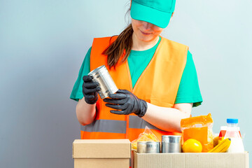 Free Food Distribution. Volunteer sorting food donation box. Young smiling woman wearing uniform cap and t-shirt, orange vest. Girl collects grocery sets, helping in-need people