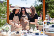 © Марина Десятниченко - Three brunette women sellers of European appearance in aprons at a street fair laughing discuss their goods