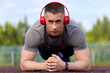 Young muscular athlete stands in the plank position, performs the exercise for a while. Strong cute athlete does push-ups outdoors to music in wireless headphones