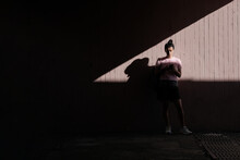 Young Woman Looking At A Phone, Standing Against Wall In Underground Pedestrian Crossing Tunnel With Contrast Shadows And Light During Sunny Summer Day. Street Style Photography