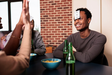 Wall Mural - Happy diverse friends playing cards game together at home while enjoying snacks and beverages. Multiethnic group of people playing guessing game with cards while having fun in living room.