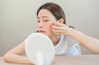 © KMPZZZ - Dermatology, puberty asian young woman, girl looking into mirror, allergy presenting an allergic reaction from cosmetic, red spot or  rash on face. Beauty care from skin problem by medical treatment.