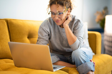 female sitting on a yellow couch using laptop and internet connection and smile. happy woman in indo