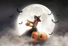 A Little Girl In A Witch Costume Sits On A Pumpkin Against The Background Of The Moon, Bats Fly On Halloween Holiday Party In Smoke