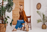 Fototapeta Boho - Fashionable woman wearing stylish boho outfit with suede jacket, wide leg jeans, sandals, holding trendy round wicker straw bag, posing in cozy room with green plants. Copy, empty space for text