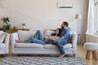 © fizkes - Happy millennial couple of homeowners enjoying cool conditioned air, resting on couch together, using remote control for AC, cooling domestic equipment start. Home appliance concept