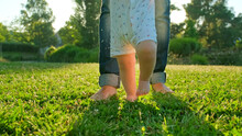 Child Learns To Take The First Steps On The Grass. Baby Learns To Walk With The Help Of His Mother On A Green Grass In The Park.  Walking Children's Bare Feet On A Green Lawn Close-up.