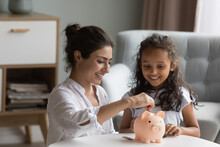 Happy Excited Indian Mother And Kid Dropping Cash Into Piggybank. Caring Mom Teaching Kid To Save, Invest Money, Collecting Coins In Piggy Bank. Family Savings, Financial Education Concept