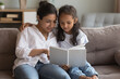 © fizkes - Happy young Indian mom teaching little daughter to read book, helping with primary school homework, showing pictures, telling fairy tale. Mother and kid girl resting on comfortable sofa