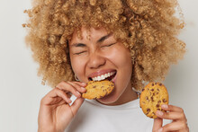 Close Up Shot Of Blonde Curly Haired Woman Eats Cookies With Chocolate Bites Deicious Snack Keeps Eyes Closed Has Sweet Tooth Dressed Casually Isolated Over White Background. Unhealthy Eating