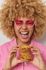 Wall Mural - Vertical shot of curly haired European woman exclaims loudly holds cookies with chocolate wears trendy sunglasses and jacket poses indoor. Optimistic female model holds delicious sweet snack