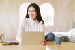 Beautiful smiling freelancer Asian woman working remotely on laptop and looking away in home winter atmosphere. Positive female entrepreneur working on new project at desk. Working from home concept.
