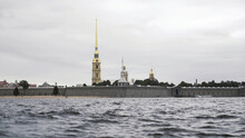 Close Up Of Small Waves Of Neva River And The Golden Spire Of The Peter And Paul Fortress. Action. Cloudy Sky Above The Petropavlovskaya Fortress And Neva River.