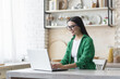 © Liubomir - Young beautiful business woman in glasses and green shirt working at home in kitchen on laptop, smiling brunette working online remotely