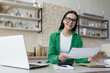 © Liubomir - Happy and successful business woman working at home in kitchen, looking at camera and smiling wearing green magpie, brunette holding letter and document