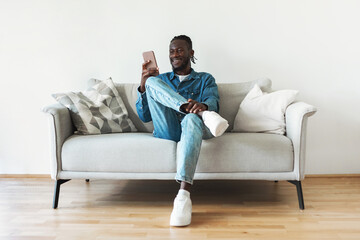 Wall Mural - African American Man Using Cellphone Texting Sitting On Couch Indoor