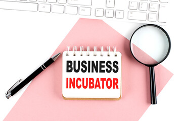 Business concept. Notebook with text BUSINESS INCUBATOR on pink paper with magnifier, keyboard and pen in the white background