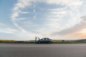 Wall Mural - A young girl stands near a broken-down car in the middle of the highway during sunset and tries to call for help on the phone. Waiting for help. Car service. Car breakdown on road.