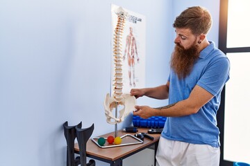 Poster - Young redhead man wearing physiotherapist uniform pointing to anatomical model of vertebral column at physiotherapy clinic