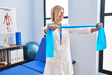 Poster - Middle age blonde woman wearing physiotherapist uniform using elastic band at physiotherapy clinic