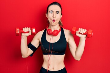 Poster - Young hispanic girl wearing sportswear using dumbbells and headphones making fish face with mouth and squinting eyes, crazy and comical.