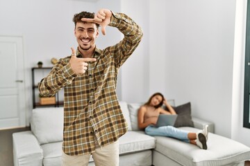 Canvas Print - Young handsome man at the living room with girlfriend smiling making frame with hands and fingers with happy face. creativity and photography concept.