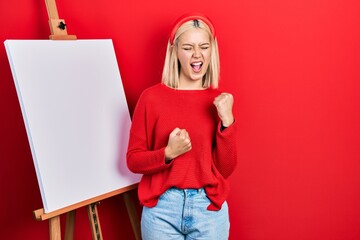 Sticker - Beautiful blonde woman standing by painter easel stand celebrating surprised and amazed for success with arms raised and eyes closed