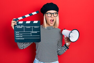 Sticker - Beautiful blonde woman holding video film clapboard and megaphone angry and mad screaming frustrated and furious, shouting with anger. rage and aggressive concept.