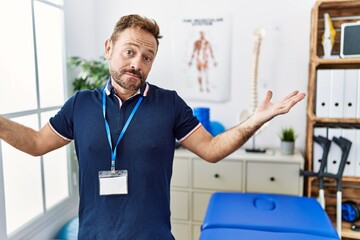 Middle age physiotherapist man working at pain recovery clinic clueless and confused expression with arms and hands raised. doubt concept.