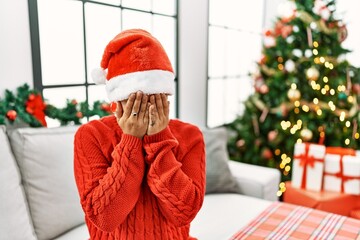 Poster - Young hispanic woman with short hair wearing christmas hat sitting on the sofa with sad expression covering face with hands while crying. depression concept.