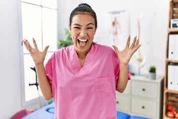 Sticker - Young hispanic woman working at pain recovery clinic celebrating crazy and amazed for success with arms raised and open eyes screaming excited. winner concept