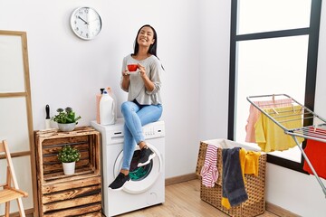 Wall Mural - Young latin woman drinking coffee waiting for washing machine at laundry room