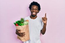 Young African American Man Holding Paper Bag With Bread And Groceries Smiling With An Idea Or Question Pointing Finger With Happy Face, Number One