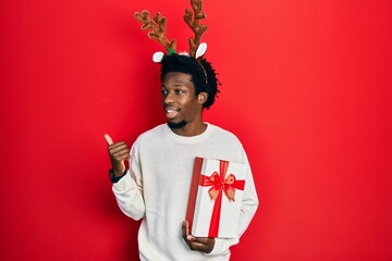 Wall Mural - Young african american man wearing deer christmas hat holding gift smiling with happy face looking and pointing to the side with thumb up.