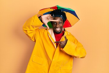 Wall Mural - Young african american man wearing yellow raincoat smiling making frame with hands and fingers with happy face. creativity and photography concept.