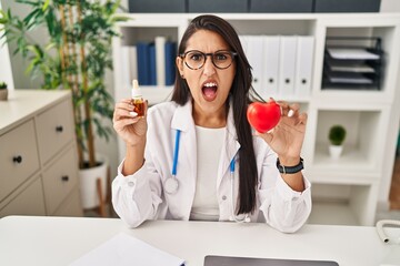 Wall Mural - Young hispanic doctor woman holding heart and cbd oil angry and mad screaming frustrated and furious, shouting with anger. rage and aggressive concept.