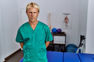 Poster - Young blond man wearing physiotherapist uniform standing at clinic skeptic and nervous, frowning upset because of problem. negative person.