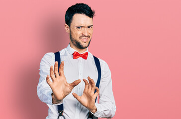 Canvas Print - Hispanic man with beard wearing hipster look with bow tie and suspenders disgusted expression, displeased and fearful doing disgust face because aversion reaction. with hands raised