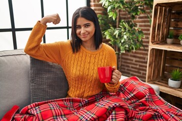 Canvas Print - Young hispanic woman sitting on the sofa drinking a coffee at home strong person showing arm muscle, confident and proud of power