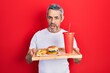Handsome middle age man with grey hair eating a tasty classic burger with fries and soda in shock face, looking skeptical and sarcastic, surprised with open mouth
