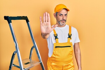 Poster - Handsome middle age man with grey hair holding ladder doing stop sing with palm of the hand. warning expression with negative and serious gesture on the face.