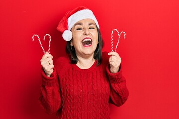 Wall Mural - Middle age hispanic woman wearing christmas hat holding candy smiling and laughing hard out loud because funny crazy joke.