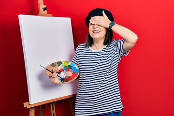 Poster - Middle age hispanic woman standing drawing with palette by painter easel stand smiling and laughing with hand on face covering eyes for surprise. blind concept.