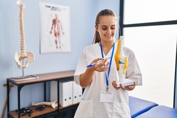 Canvas Print - Young blonde woman wearing physiotherapy uniform holding anatomical model of knee at clinic