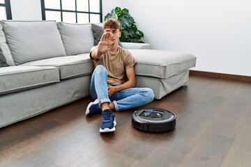 Canvas Print - Young caucasian man sitting at home by vacuum robot with open hand doing stop sign with serious and confident expression, defense gesture